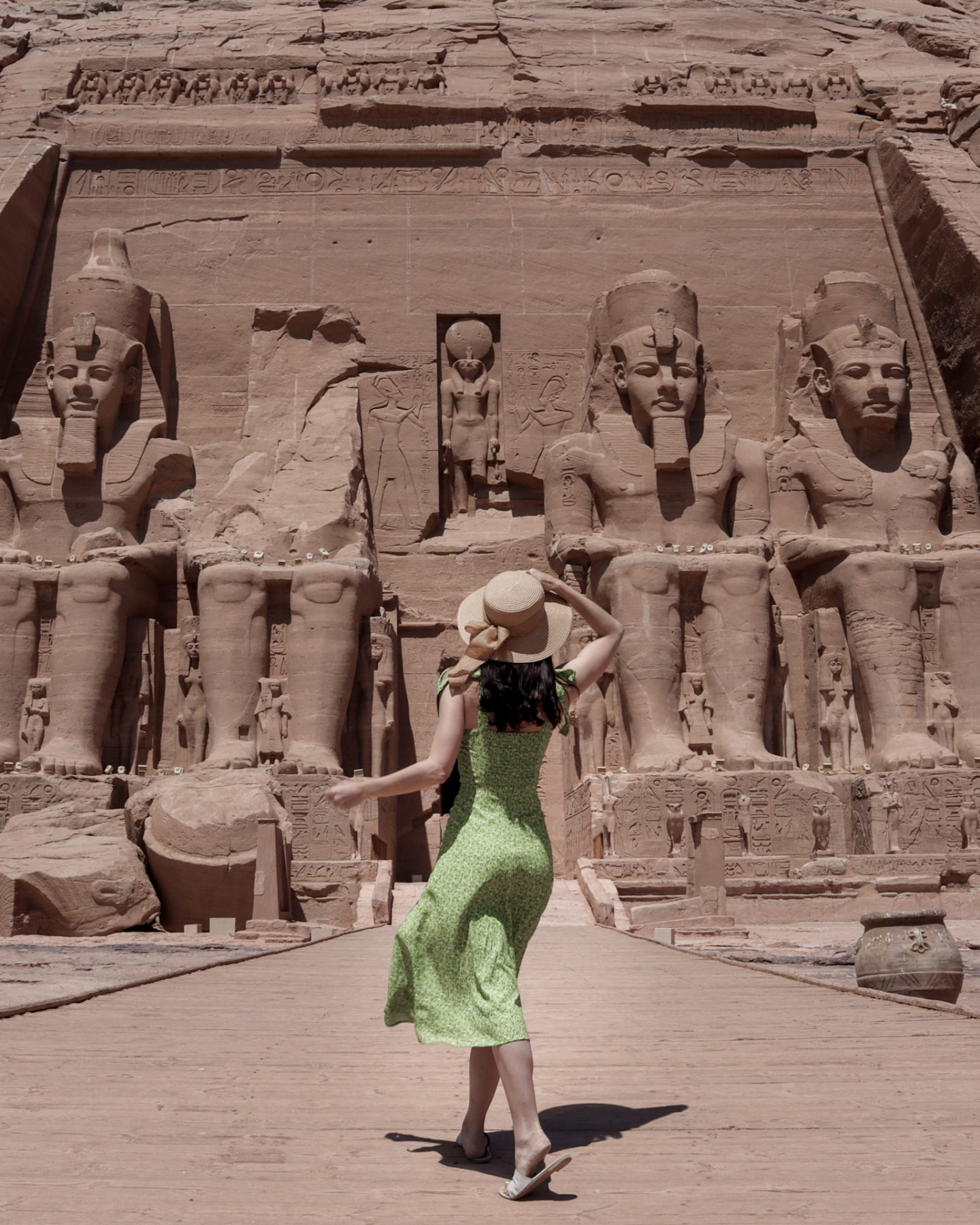 Travel Blogger Jordan Gassner wearing a green dress and with a hand holding a sunhat on her head while looking up in awe and away from the camera on the walkway leading up to the four massive sitting statues of Ramses II at Abu Simbel Temple in Egypt