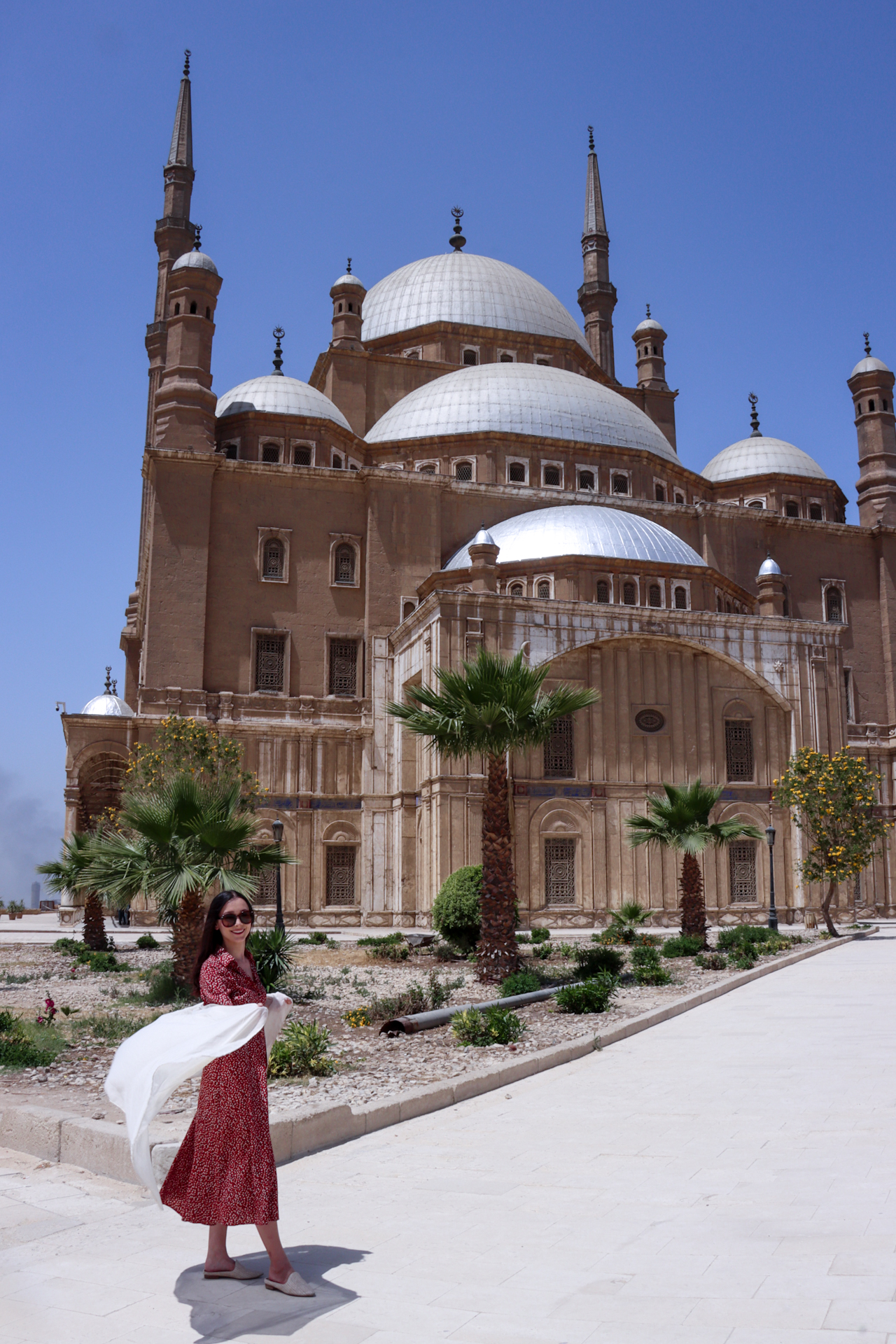 What to Wear in Egypt: Brunette woman wearing a cream scarf, brown sunglasses, a red midi shirt dress, and cream slip-on shoes in front of Muhammad Ali Mosque in Cairo, Egypt