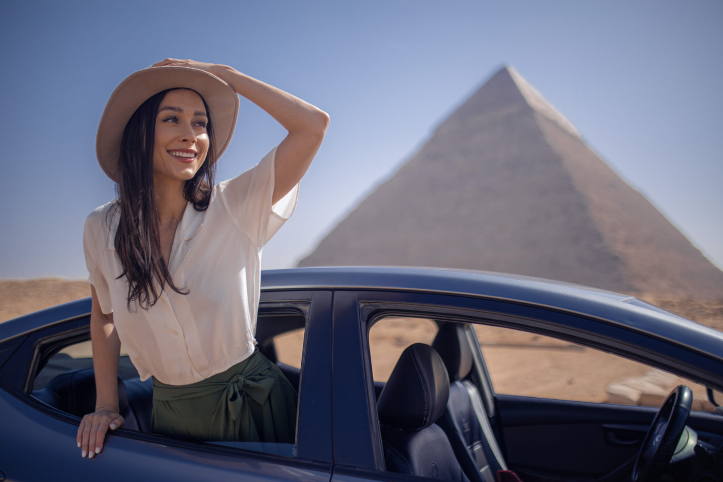 Jordan Gassner holding her explorer hat while standing up through the open window of a car in front of the Great Pyramid on the Giza Plateau in Egypt