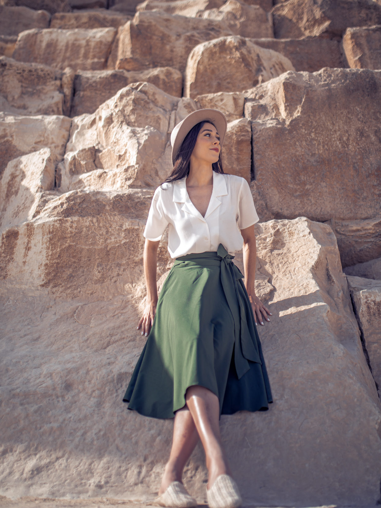 Jordan Gassner wearing an explorer hat, white button up shirt, a green midi skirt and straw mules while leaning against one of the three Giza pyramids in Egypt