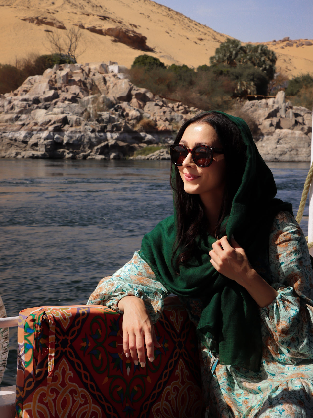 Jordan Gassner wearing round sunglasses, a turquoise dress with long sleeves, and a hunter green head scarf while sitting against a colorful seat cushion on a Nile Cruise Boat. 