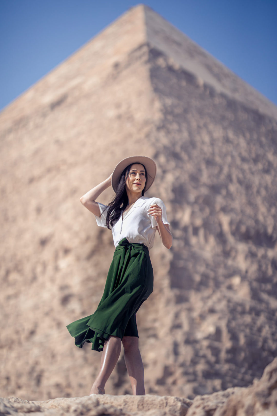 Jordan Gassner clutching her explorer hat while wearing an Indiana Jones inspired travel outfit in front of one of the Pyramids at the Giza Plateau in Egypt