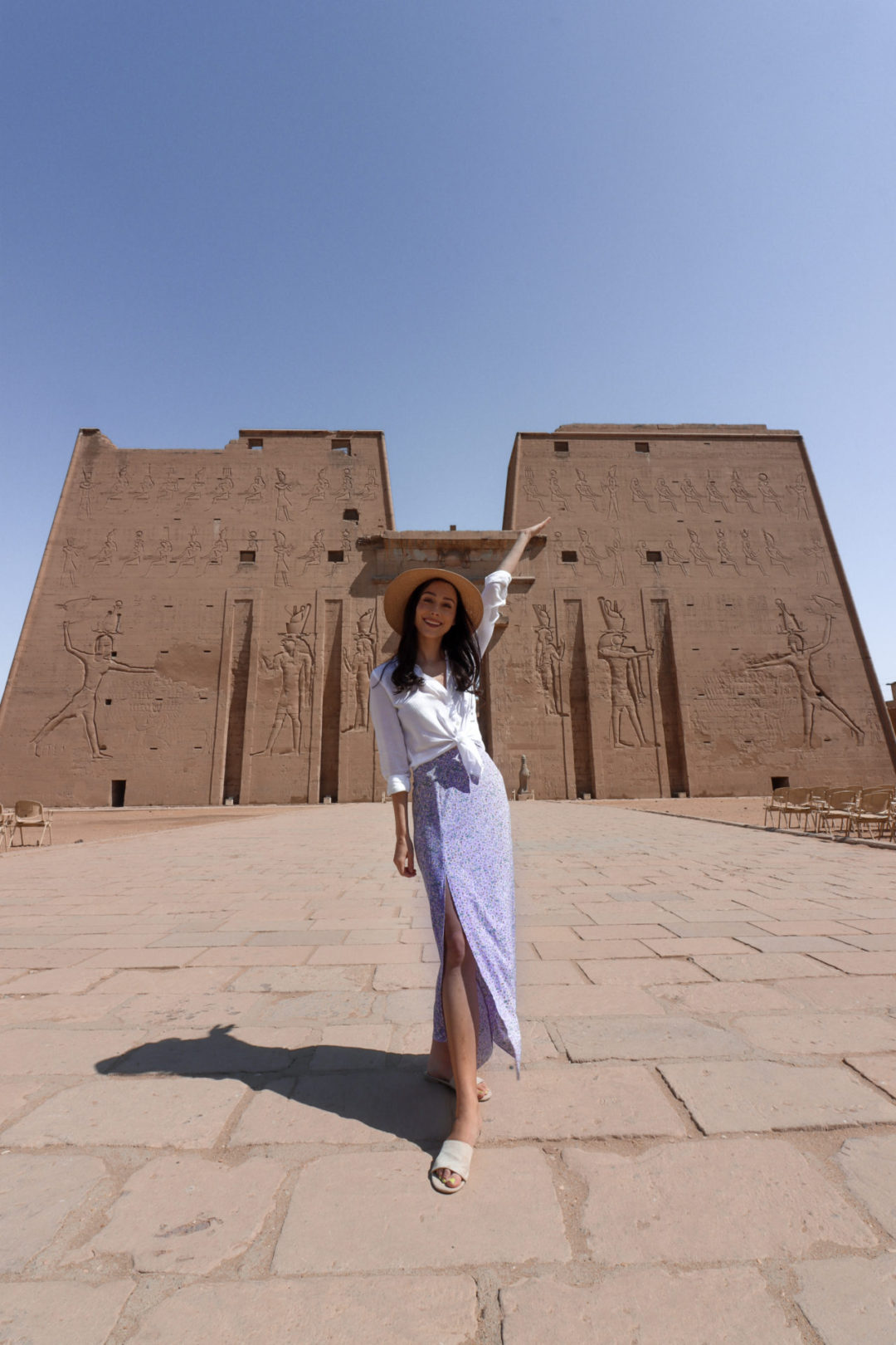 What to Wear in Egypt: Female Traveler with long brown hair wearing a straw sunhat, a white 3/4-length sleeve white shirt, purple skirt and cream sandals standing smiling in front of Edfu Temple in Egypt