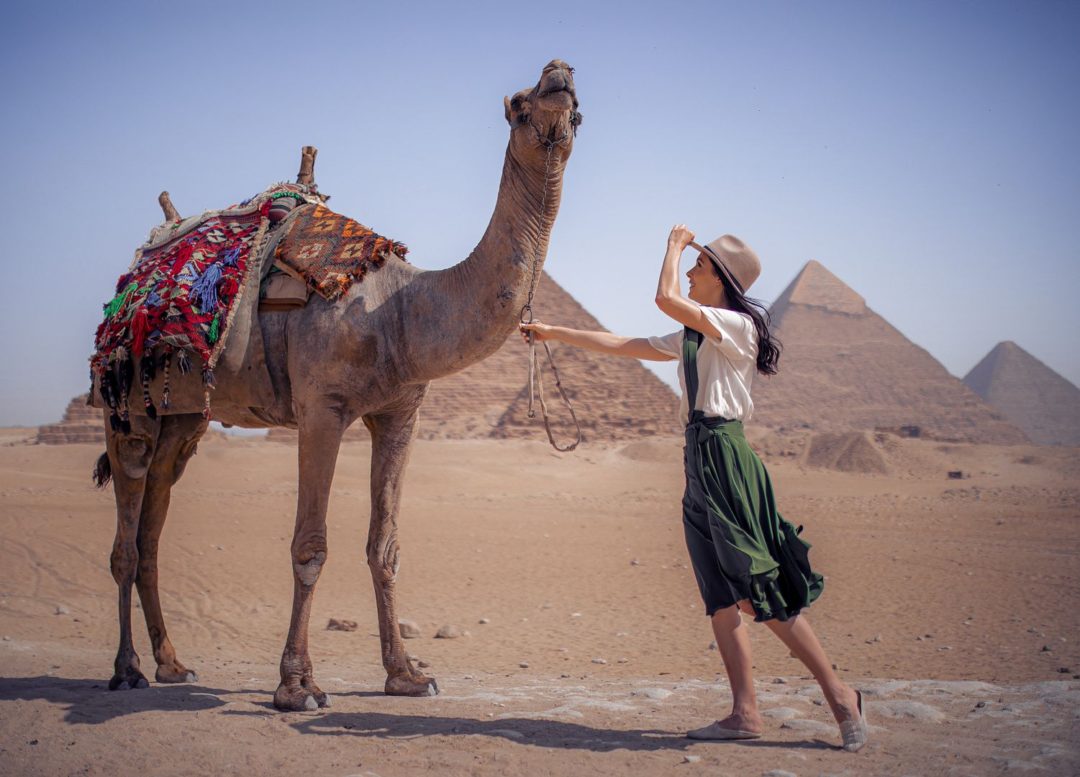Jordan Gassner holding onto a camel and her explorer hat in front of the Pyramids on the windy Giza Plateau in Egypt