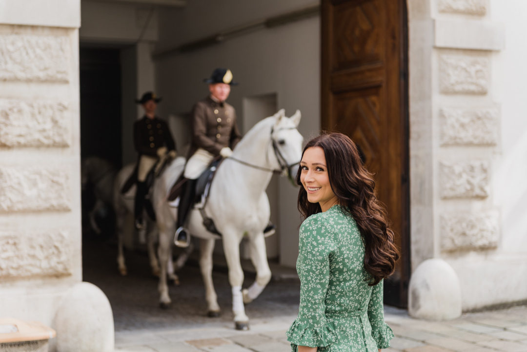 Jordan Gassner smiling over her shoulder as the Spanish Riding School passes behind her on the streets of Vienna, Austria