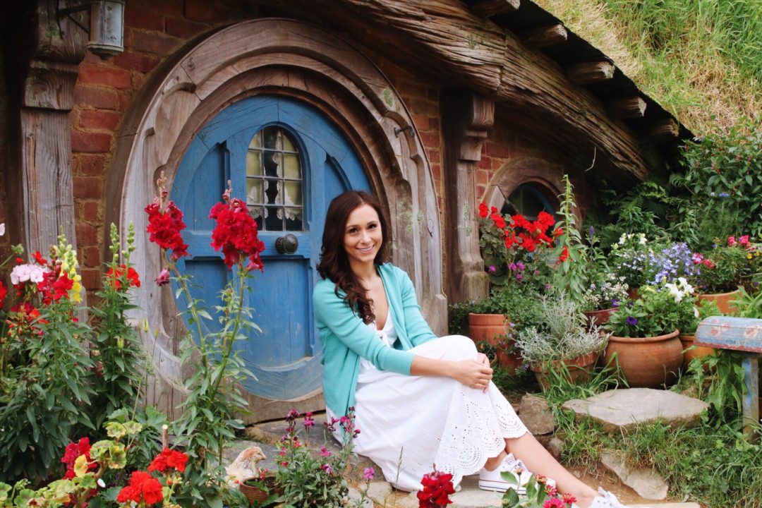 Visit New Zealand: Travel Blogger, Jordan Gassner wearing a white dress and turquoise cardigan while sitting in front of a blue round door of a Hobbit House in Hobbiton, New Zealand