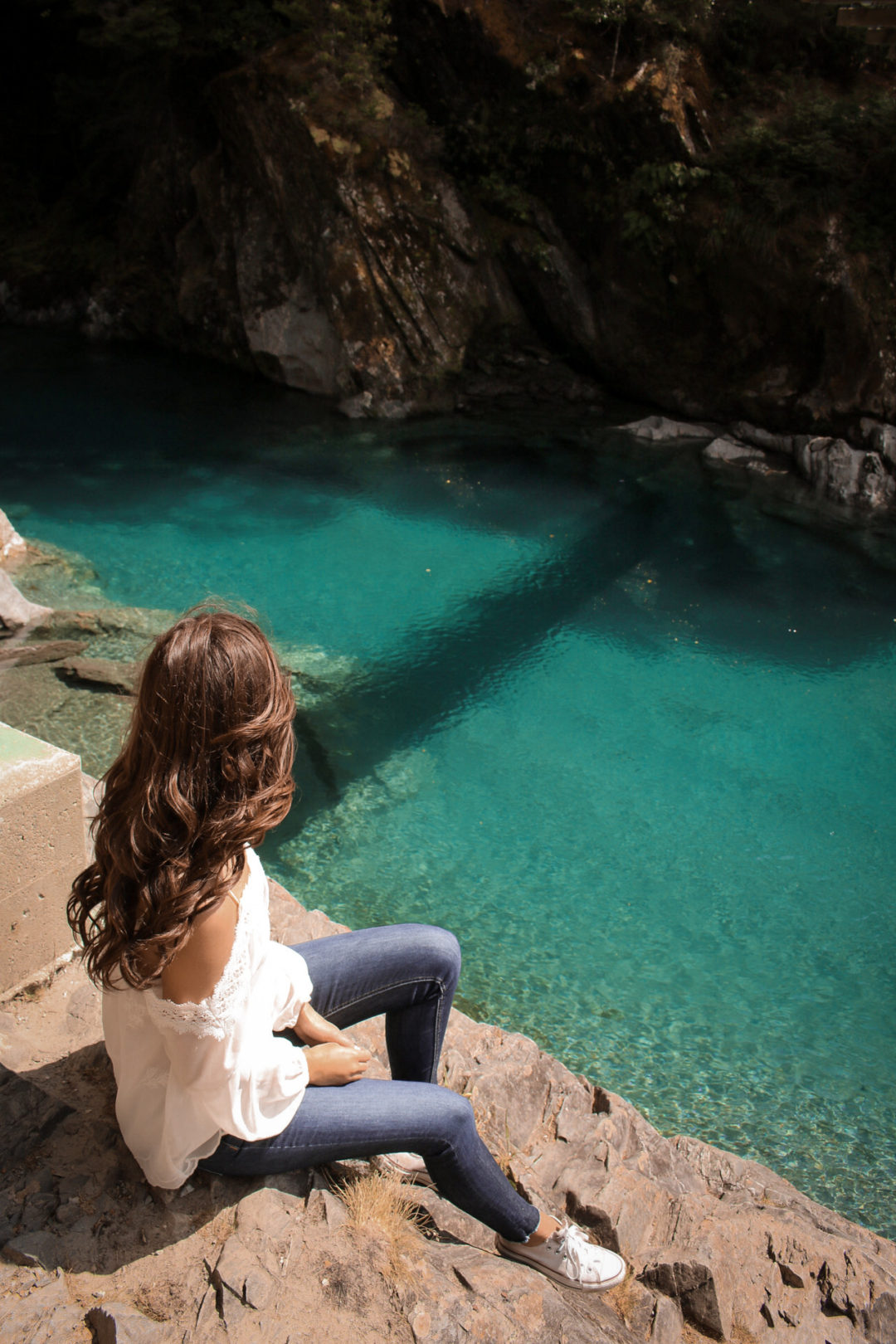 Visiting New Zealand: Brunette female blogger in white long sleeve, off-shoulder top, skinny jeans and white converse sitting on the rocks along along a glacial pool
