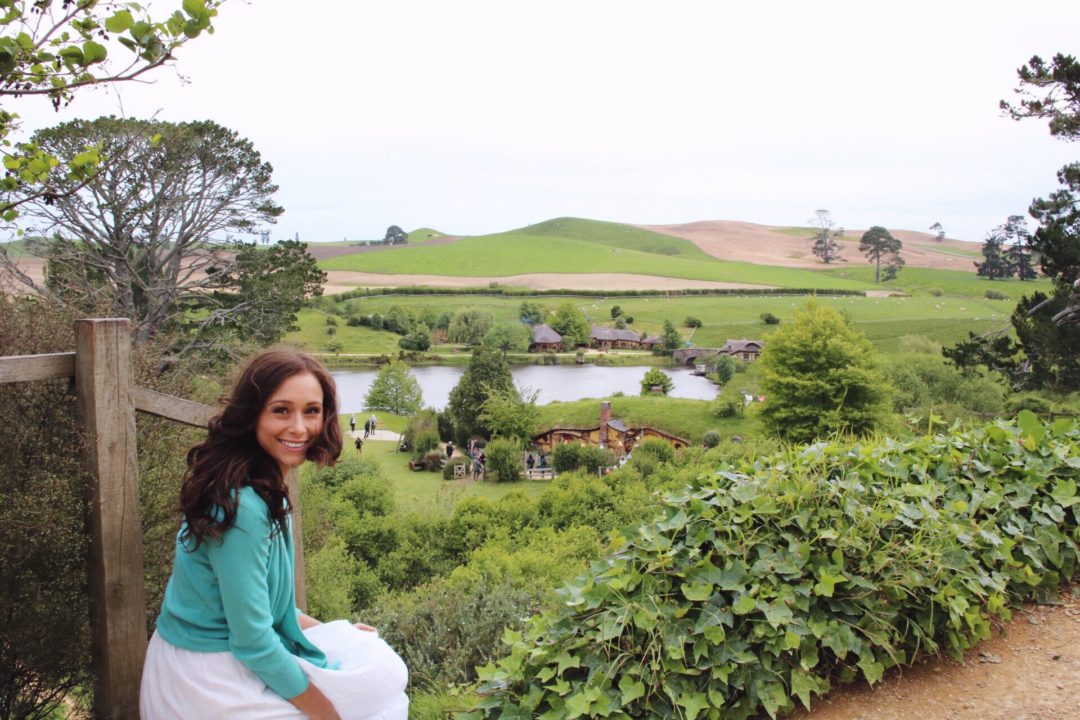 Visit New Zealand: Travel Blogger, Jordan Gassner smiling through the wind while looking back over the lookout of the Party Field and Green Dragon Pub in Hobbiton, New Zealand