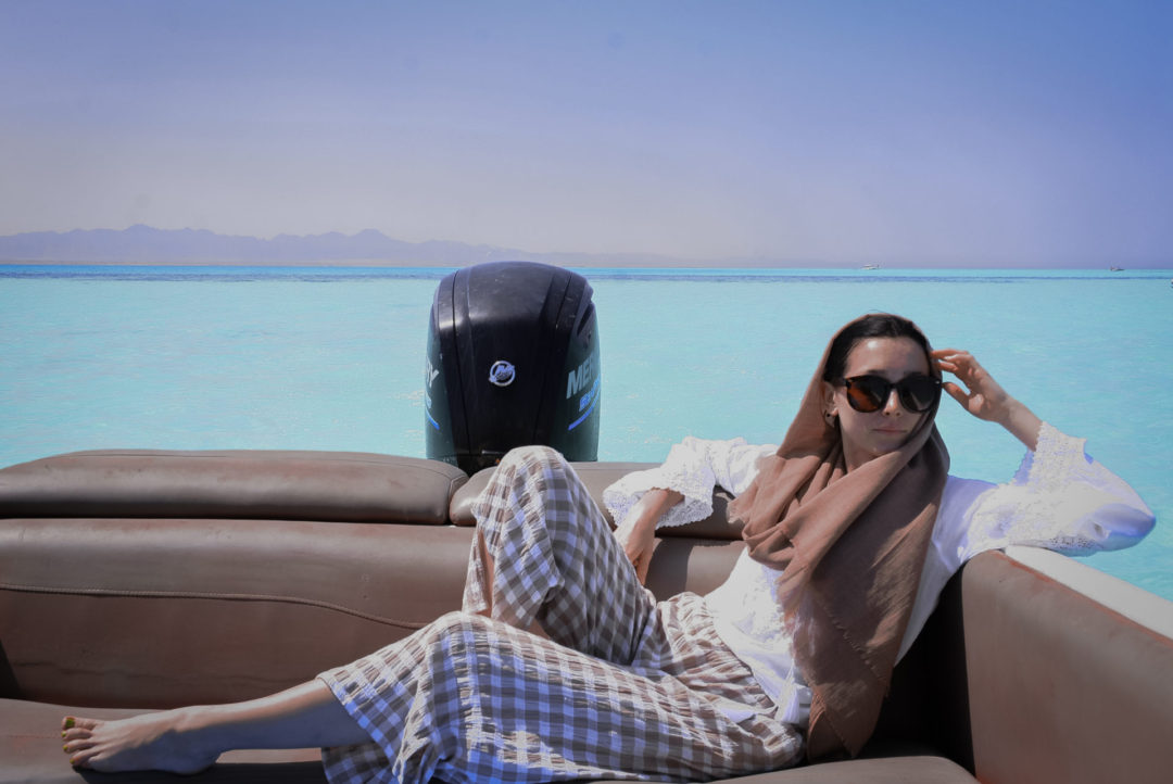 Jordan Gassner wearing a light brown head scarf, round sunglasses, a white breezy top and brown and white checkerboard-patterned fabric pants while lounging in a boat in the turquoise waters of the Red Sea off the coast of Egypt