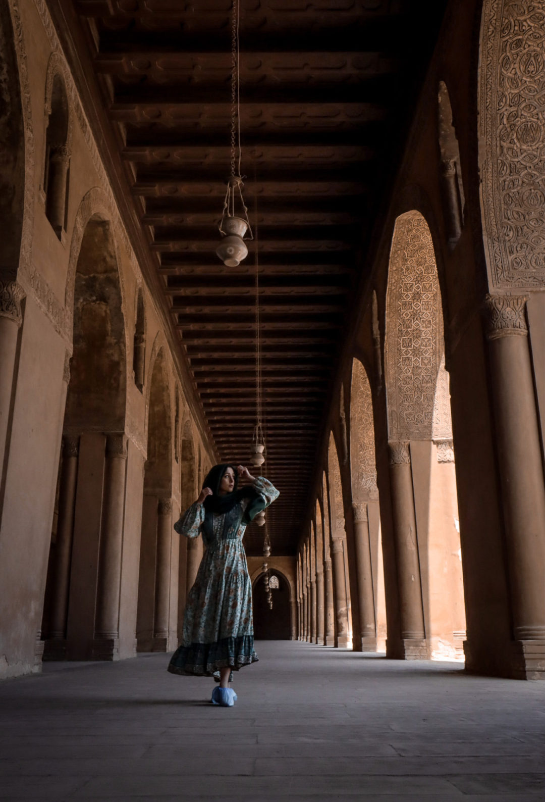 Jordan Gassner walking in a turquoise dress, hunter green head scarf and slip-on disposable socks throughout one of the arcades inside the one of the must-see Cairo Mosques: the Mosque of Ibn Tulun