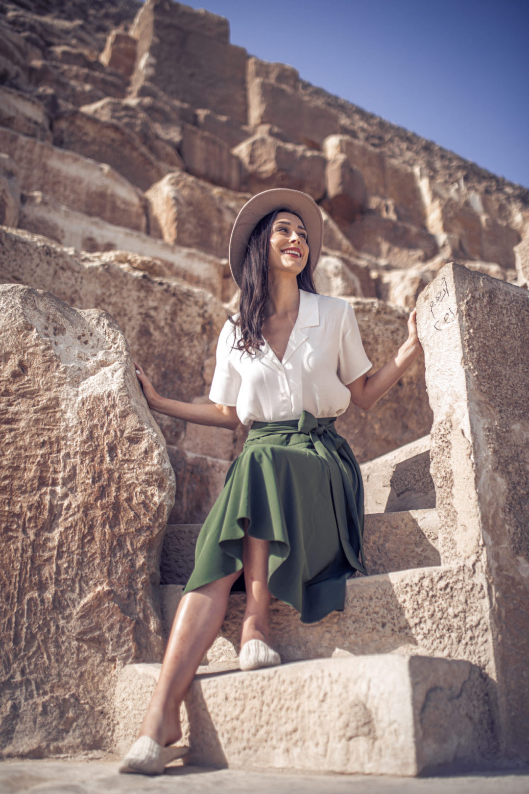 Travel Blogger Jordan Gassner wearing an explorer hat, white button up shirt, a green midi skirt and straw mules while leaning against one of the three Giza pyramids in Egypt