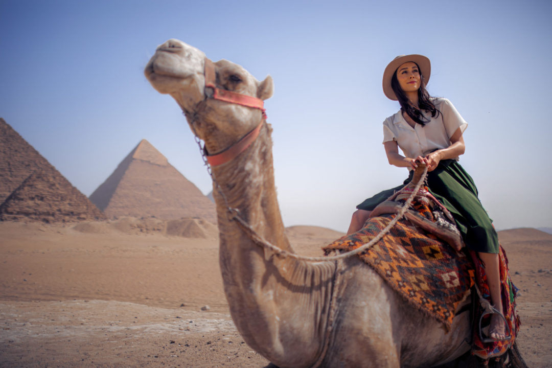 Jordan Gassner riding a camel in a vintage-inspired explorer outfit in front of the Pyramids on the windy Giza Plateau in Egypt