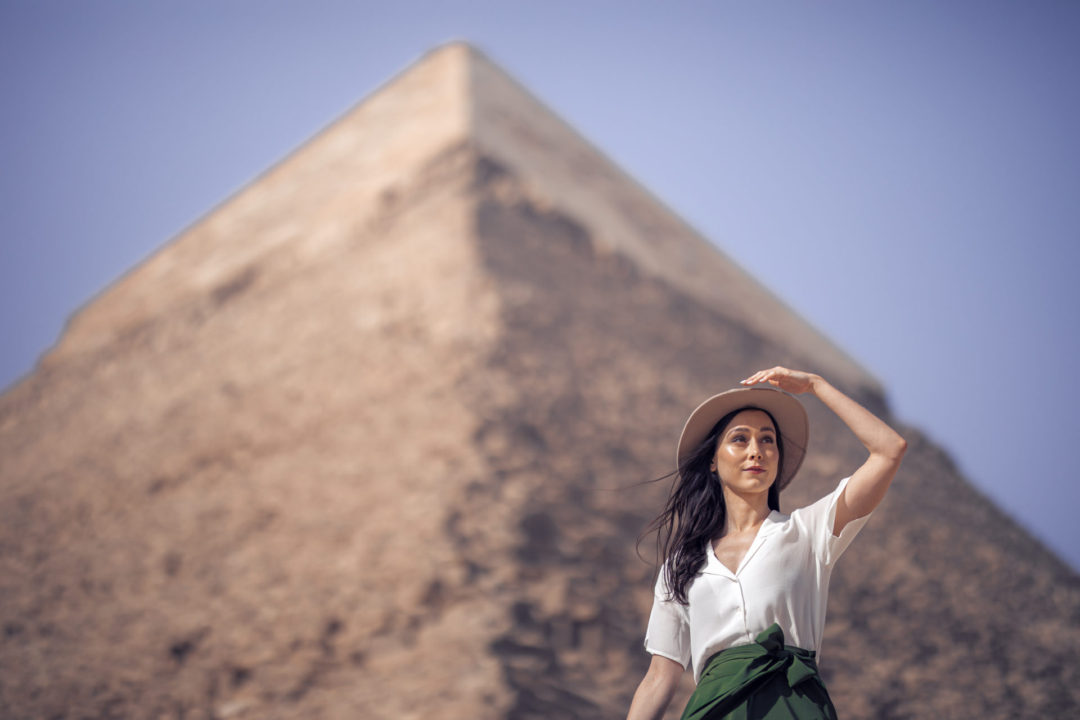 Jordan Gassner shielding her eyes from the sun and wearing a vintage-inspired explorer outfit in front of one of the pyramids at the Giza Plateau in Egypt