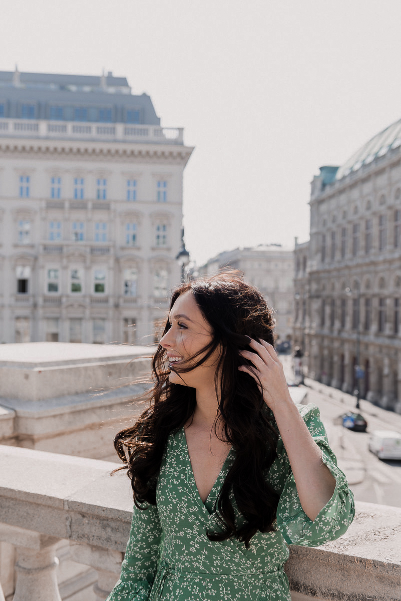 Jordan Gassner wearing a green floral dress and fighting the wind on top of a balcony in front of Vienna's Opera House in Austria