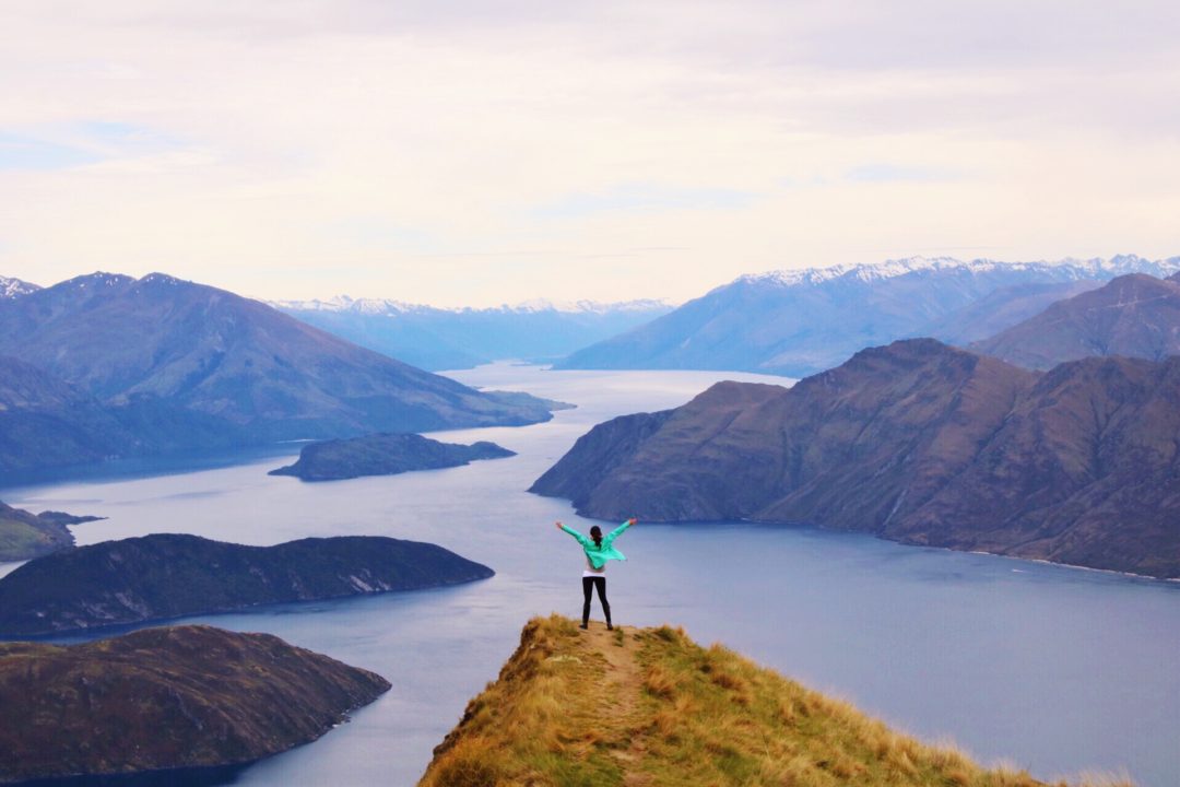 Visit New Zealand: Travel Blogger Jordan Gassner wearing a bright turqouise rain coat standing with arms triumphantly spread overhead on top of Roy's Peak Lookout over Lake Wanaka in New Zealand