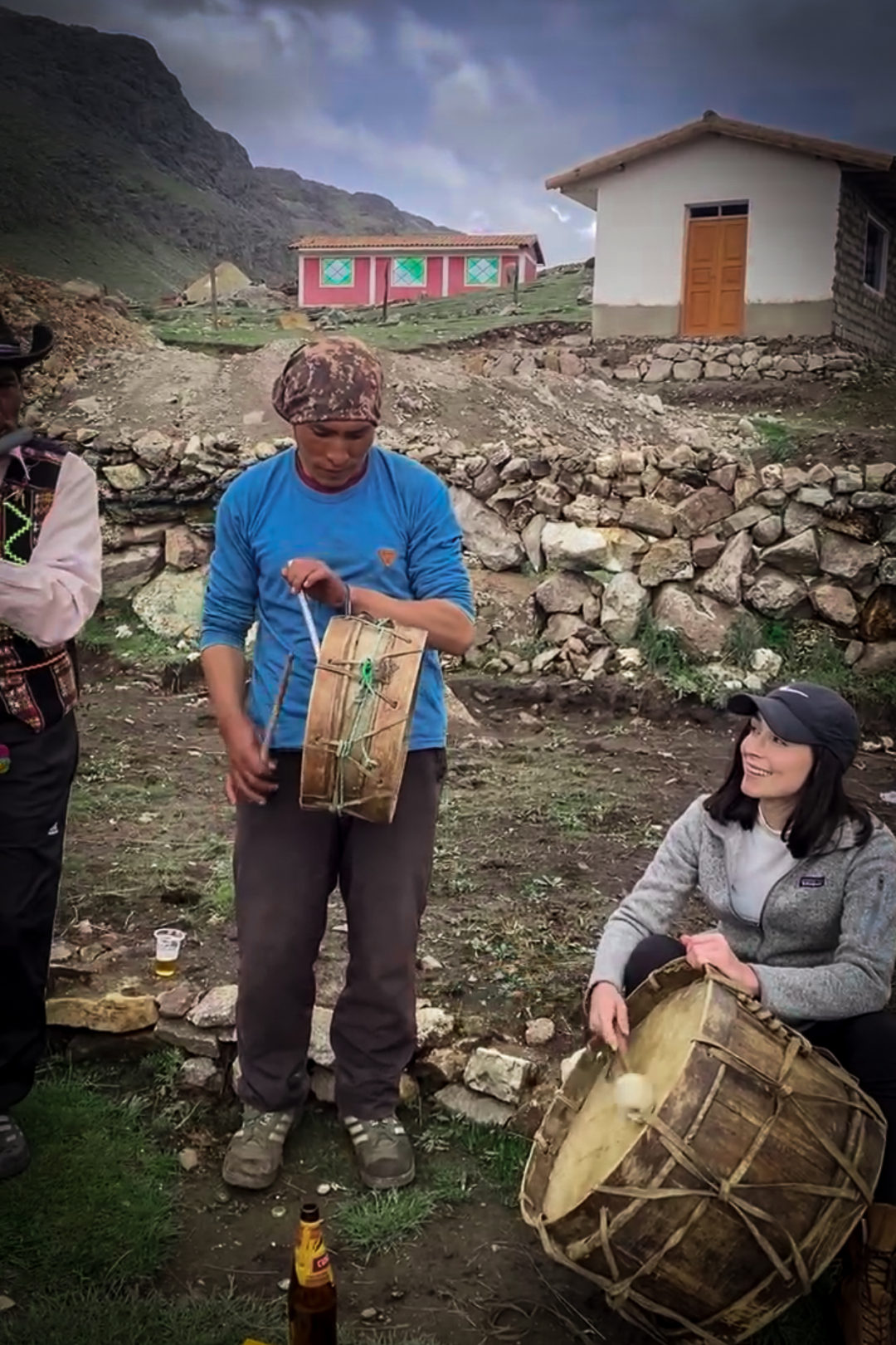 Local Quechua musician playing a harmonica, Local Quechua Chef wearing a blue shirt and playing a small drum next to Travel Blogger Jordan Gassner sitting and wearing a grey Patagonia zip-up sweatshirt while playing a large traditional Quechua drum 