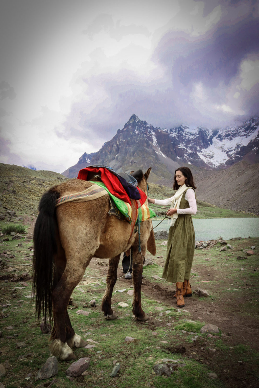 Travel Blogger Jordan Gassner wearing a dark green fantasy inspired maxi dress, white long sleeve shirt, cream alpaca wool scarf and brown hiking boots while holding the reigns and petting a small buckskin-coated horse with a colorful saddle and riding accessories in front of a glacial lake on the Ausangate 7 Lakes Trek in the Peruvian Andes