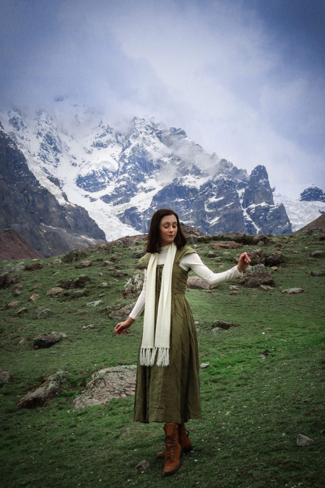 Travel Blogger Jordan Gassner looking down with her arms out while standing on a hill and wearing a dark green fantasy inspired maxi dress, white long sleeve shirt, cream alpaca wool scarf and brown hiking boots on the Ausangate 7 Lakes Trek near Cusco, Peru