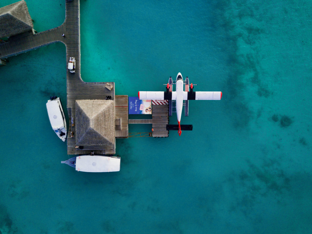 Experiences in Ambergris Caye: Drone view of a seaplane pulled up to a dock with a palapa hut in turquoise ocean waters