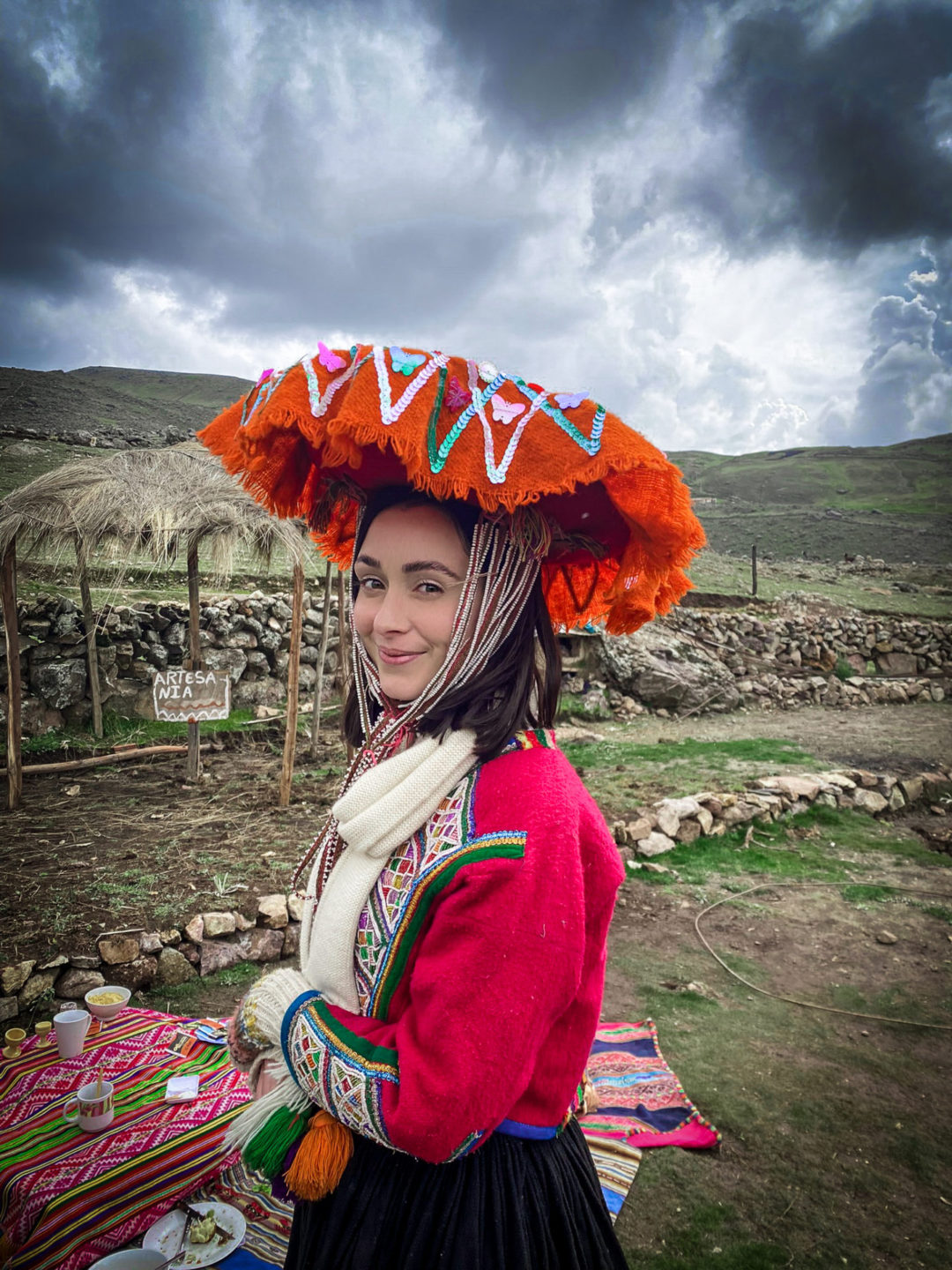 Female Travel Blogger smiling at-camera and wearing a traditional Quechua skirt, jacket and hat in the remote Peruvian Andes at a local picnic 