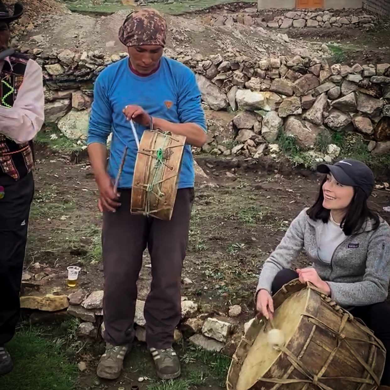 Local Quechua musician playing a harmonica, Local Quechua Chef wearing a blue shirt and playing a small drum next to Travel Blogger Jordan Gassner sitting and wearing a grey Patagonia zip-up sweatshirt while playing a large traditional Quechua drum 
