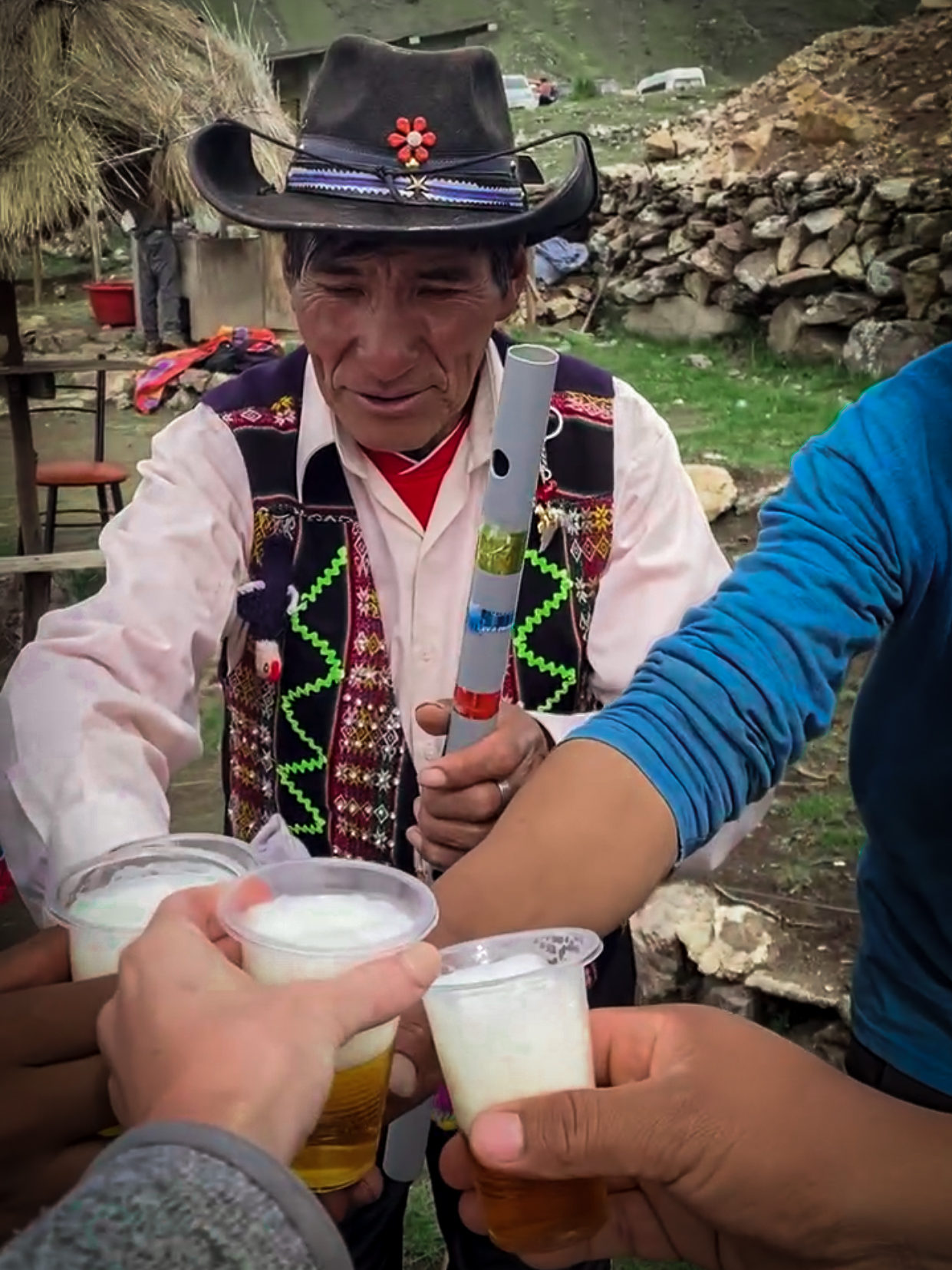 Local Quechua musician with a cowboy hat and a traditional vest holding a flute and cheers-ing four others holding clear plastic glasses of beer.