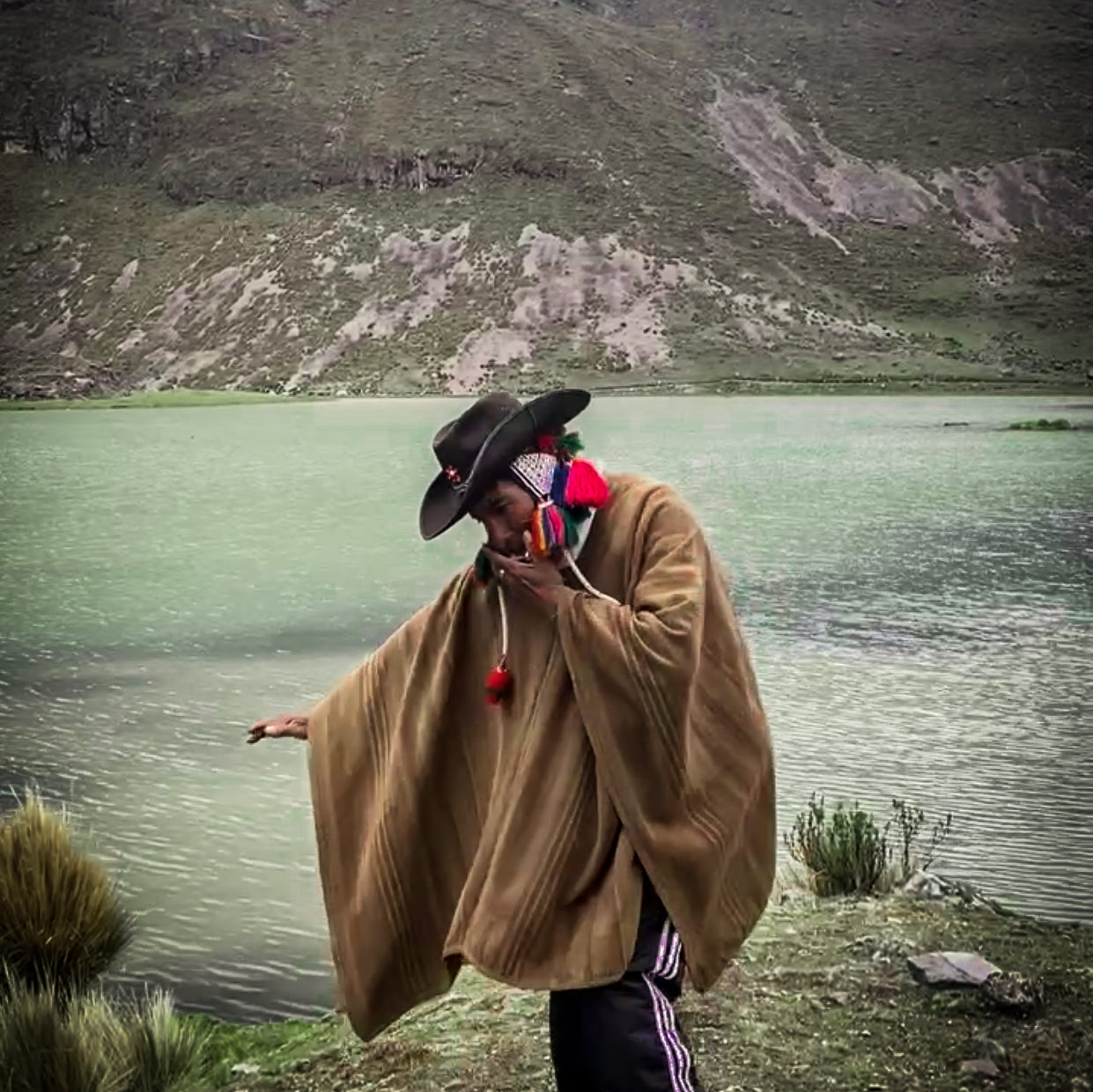 Local Quechua musician playing his harmonica and wearing a traditional poncho over Adidas pants and rainbow Quechua knit hat under a cowboy hat in front of a glacial lake and mountain landscape on the Ausangate 7 Lakes Trail in the Peruvian Andes