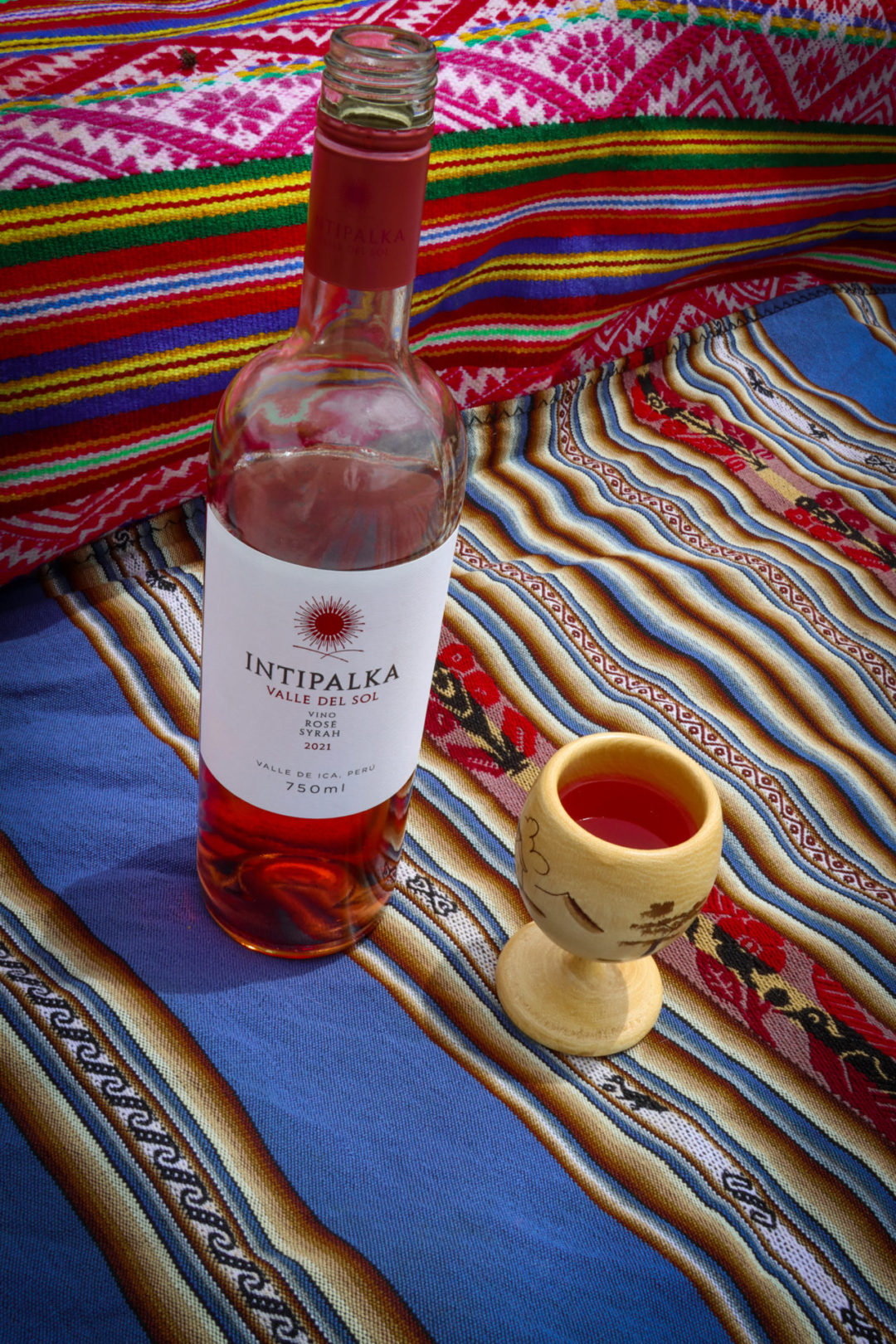 An open bottle of Intipalka Rose Wine  standing on a pink, blue, yellow and red striped Peruvian blanket next to a small yellow-painted wooden glass holding rose wine. 