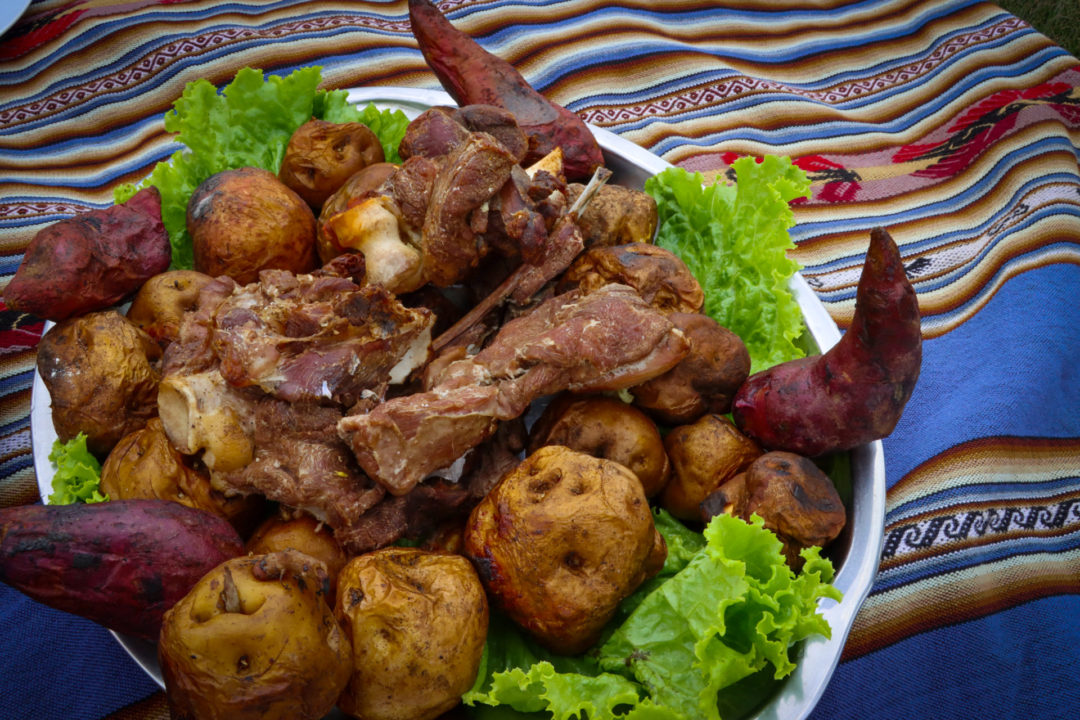 A Pachamanca tray of alpaca meat and potatoes that had been buried and cooked in the Earth served on a bed of lettuce and silver tray laying on a colorful striped Peruvian blanket