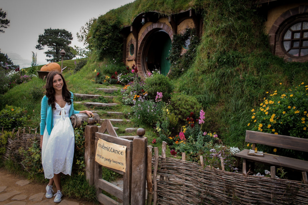 Visiting New Zealand: Travel Blogger Jordan Gassner wearing a white dress, turquoise cardigan and white converse and leaning up against the mailbox and "No Admittance except on Party Business"-posted gate at Bag End in the Hobbiton Movie Set at Matamata, New Zealand