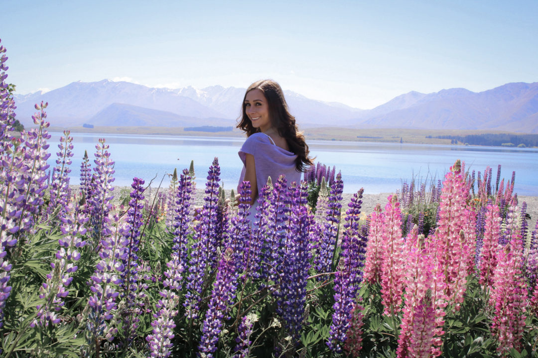 Visit New Zealand: Travel Blogger, Jordan Gassner wearing a lavender sweater and smiling back in a field of purple and pink lupins just offshore Lake Tekapo in New Zealand