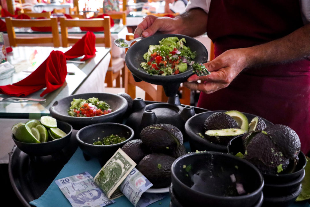 Man lifting up a goblet of guacamole with pico de gallo, lime and jalapeno from a guacamole cart with black bowls full of ingredients and Mexican pesos.