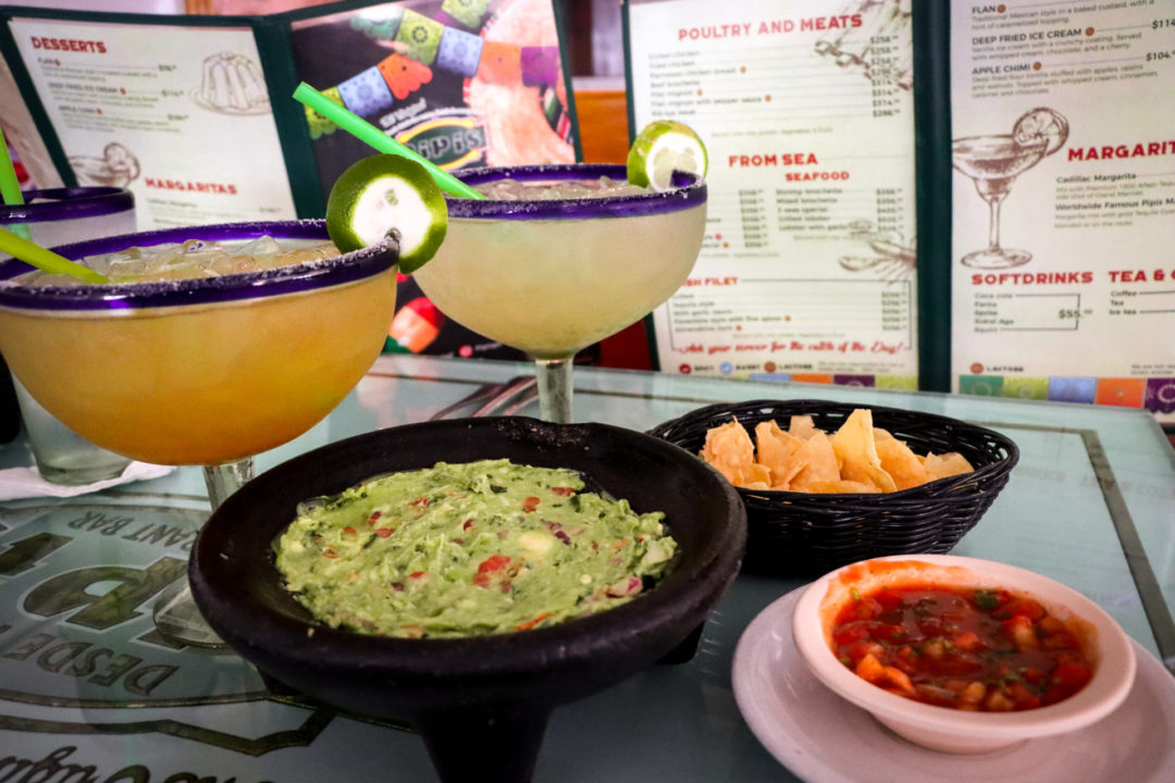 Two large margaritas in clear glasses with limes and colorful straws sit near chips, guacamole and salsa