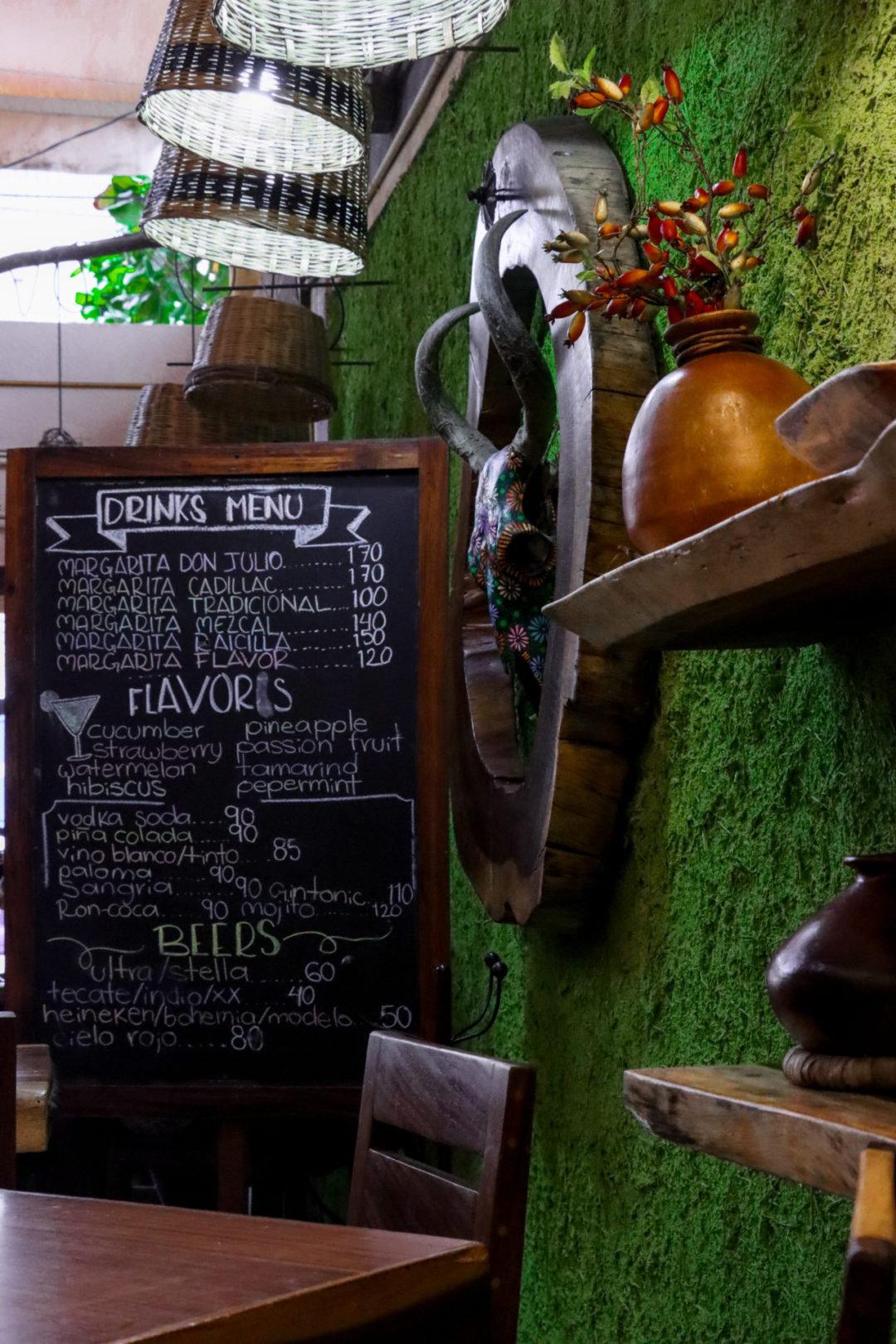 A blackboard with a wooden frame reading "Drinks menu" stands next to a grass wall with wooden decorations and a hand painted cow skull hanging up.