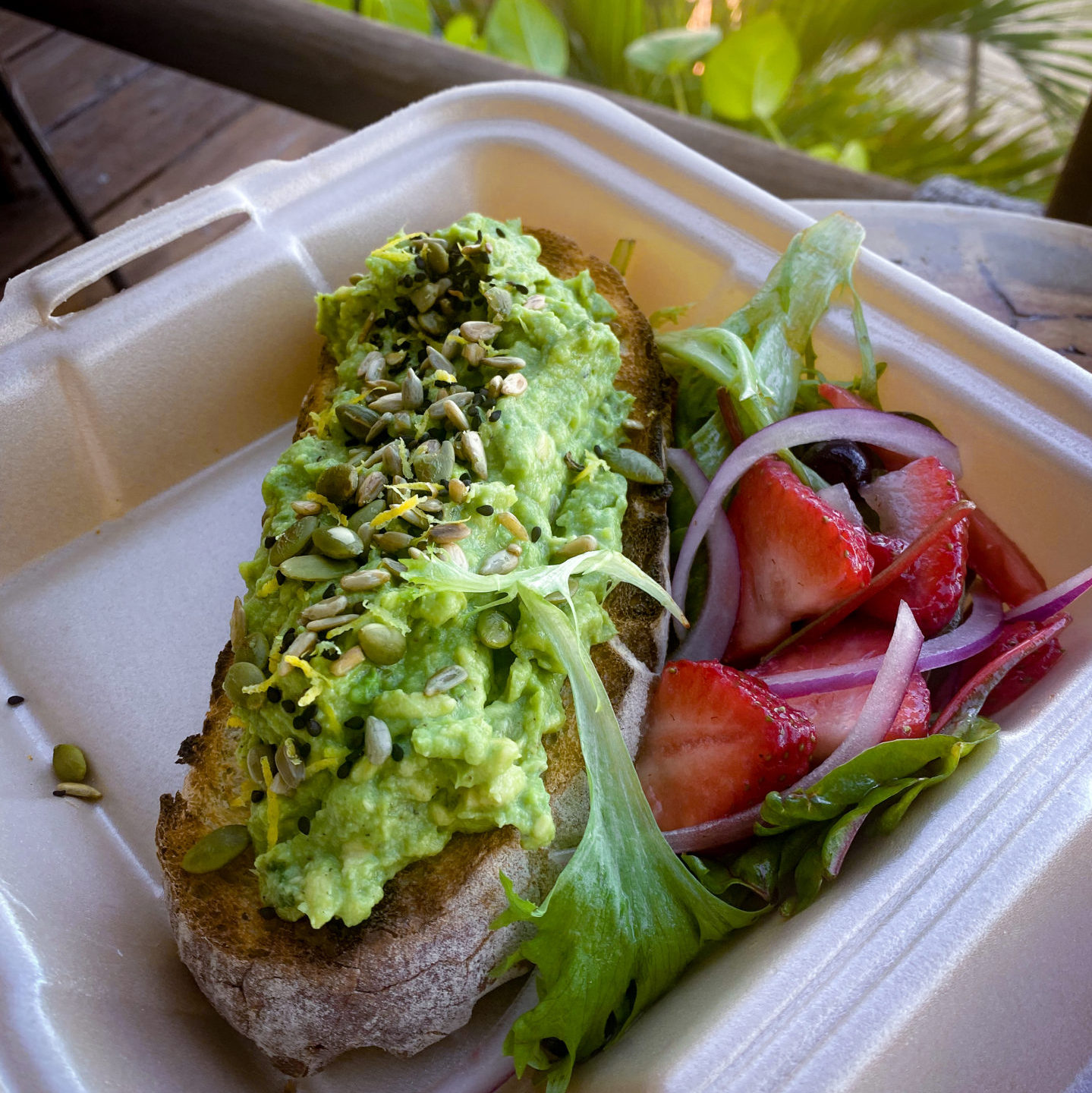 Avocado toast with orange zest and seeds next to a bed of lettuce, onion and strawberry slices in a to go box