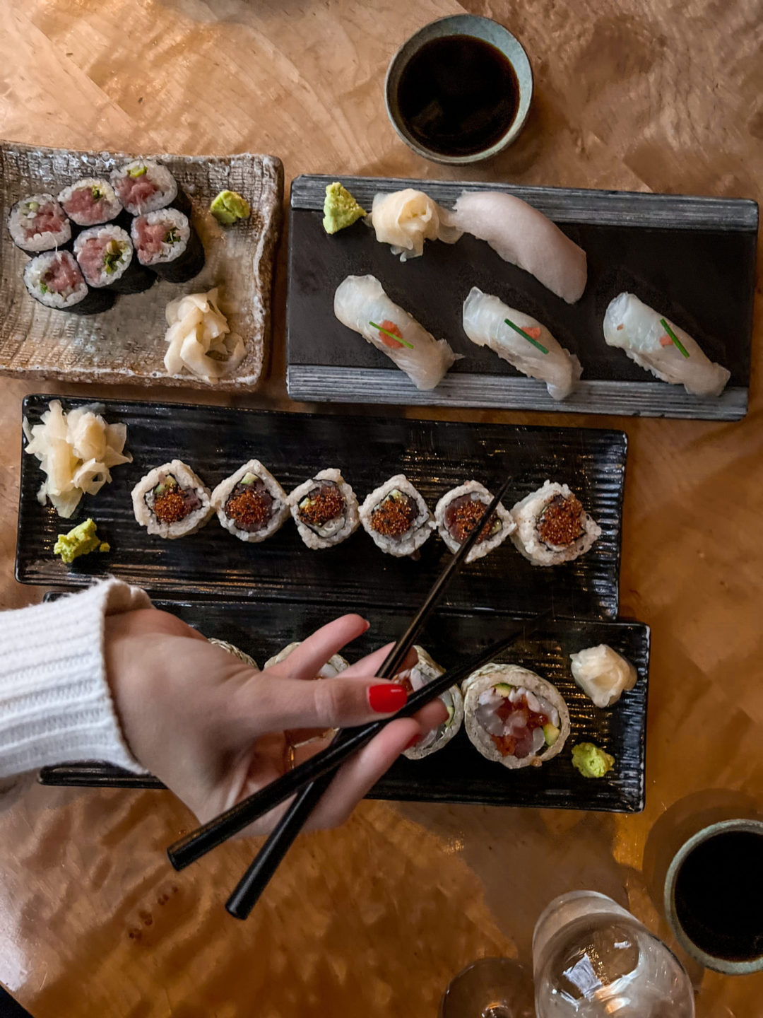 A woman's hand with red nail polish holding chopsticks over four plates of sushi from Sushi Ran in Sausalito, Marin County, California