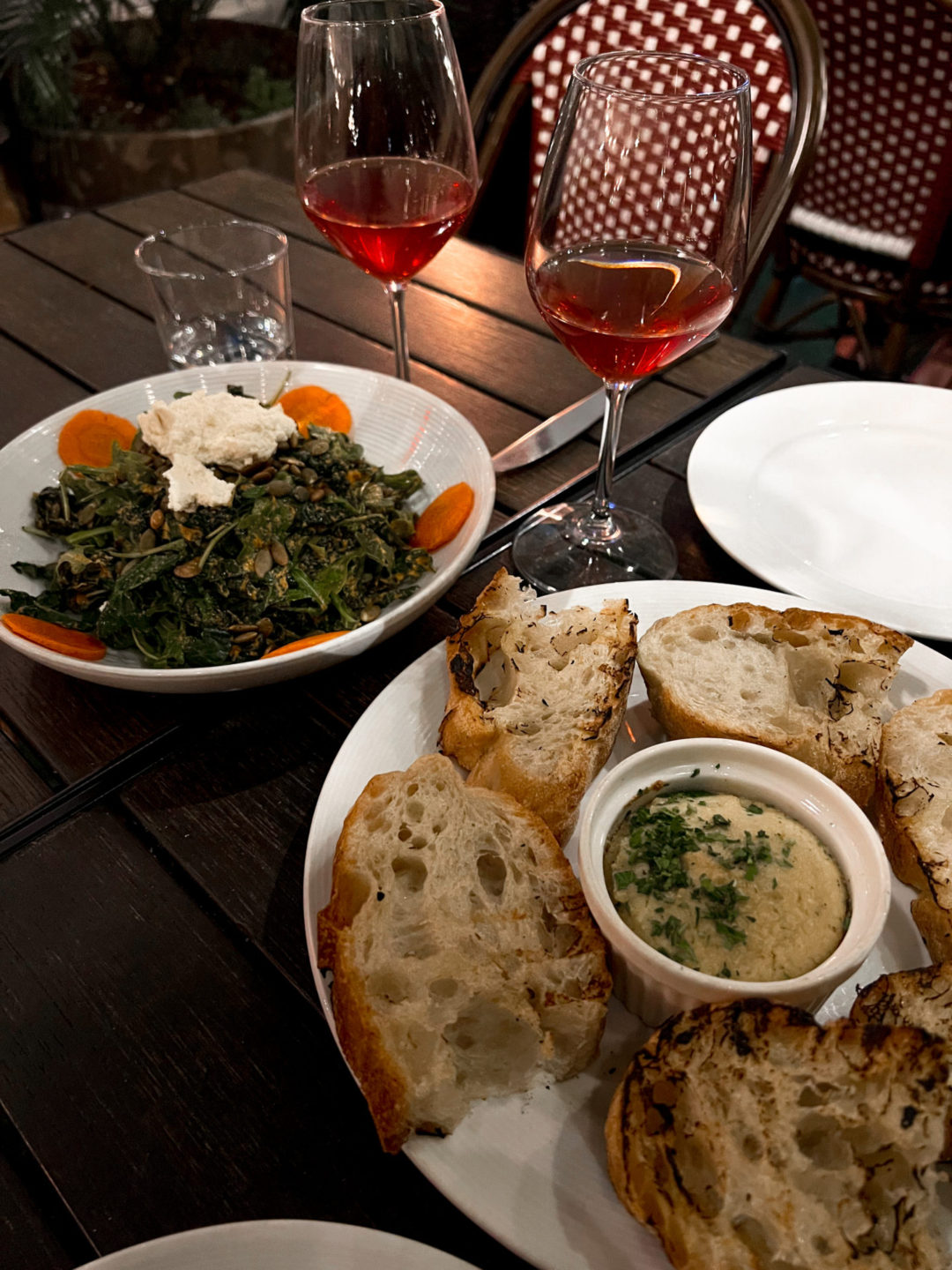 "Pura Vita" Kale Salad, Grilled Ciabatta and Cashew Dip and Two Glasses of Natural Red Wine on top of a brown table near a red and white whicker chair at Pura Vita Restaurant in West Hollywood, California