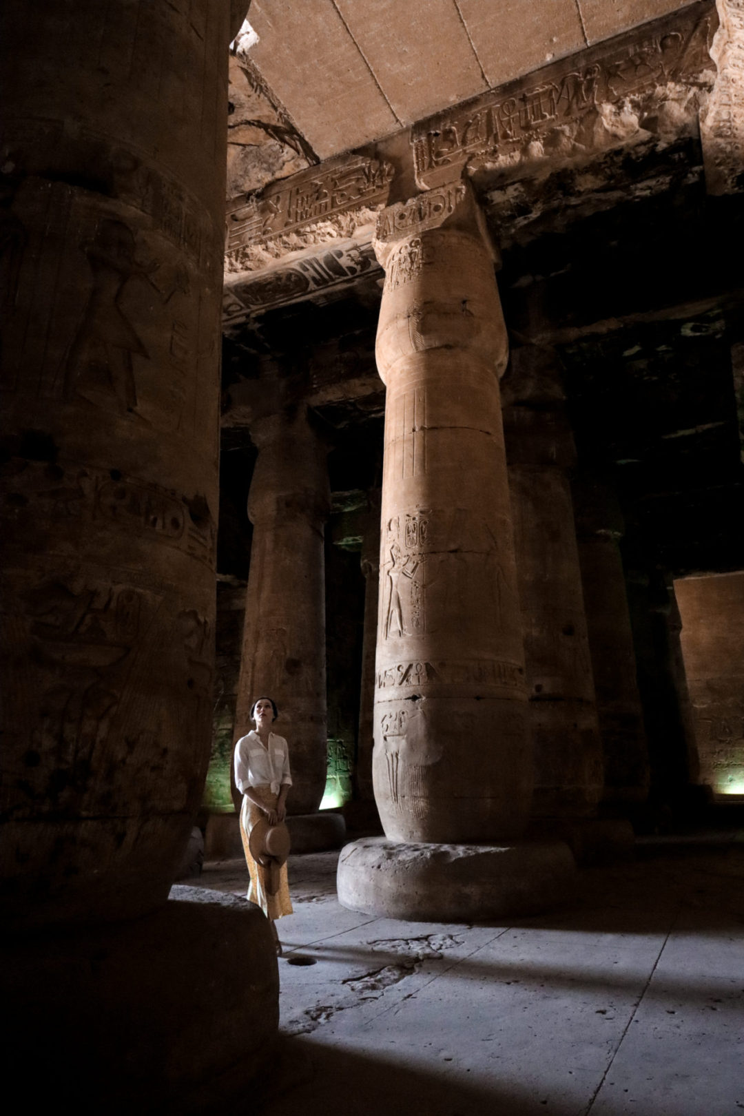 Travel Blogger Jordan Gassner wearing a yellow skirt, white button up shirt and straw hat walking into the dark, covered Abydos Temple ruins.