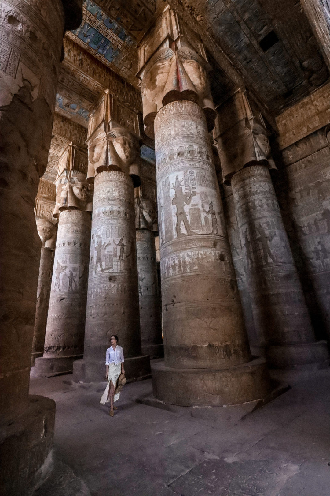 Travel Blogger Jordan Gassner wearing a white button down top and light yellow skirt with a leg slit walking next to the gigantic columns inside Dendera Temple's blue-ceilinged Hypostyle Hall in Egypt