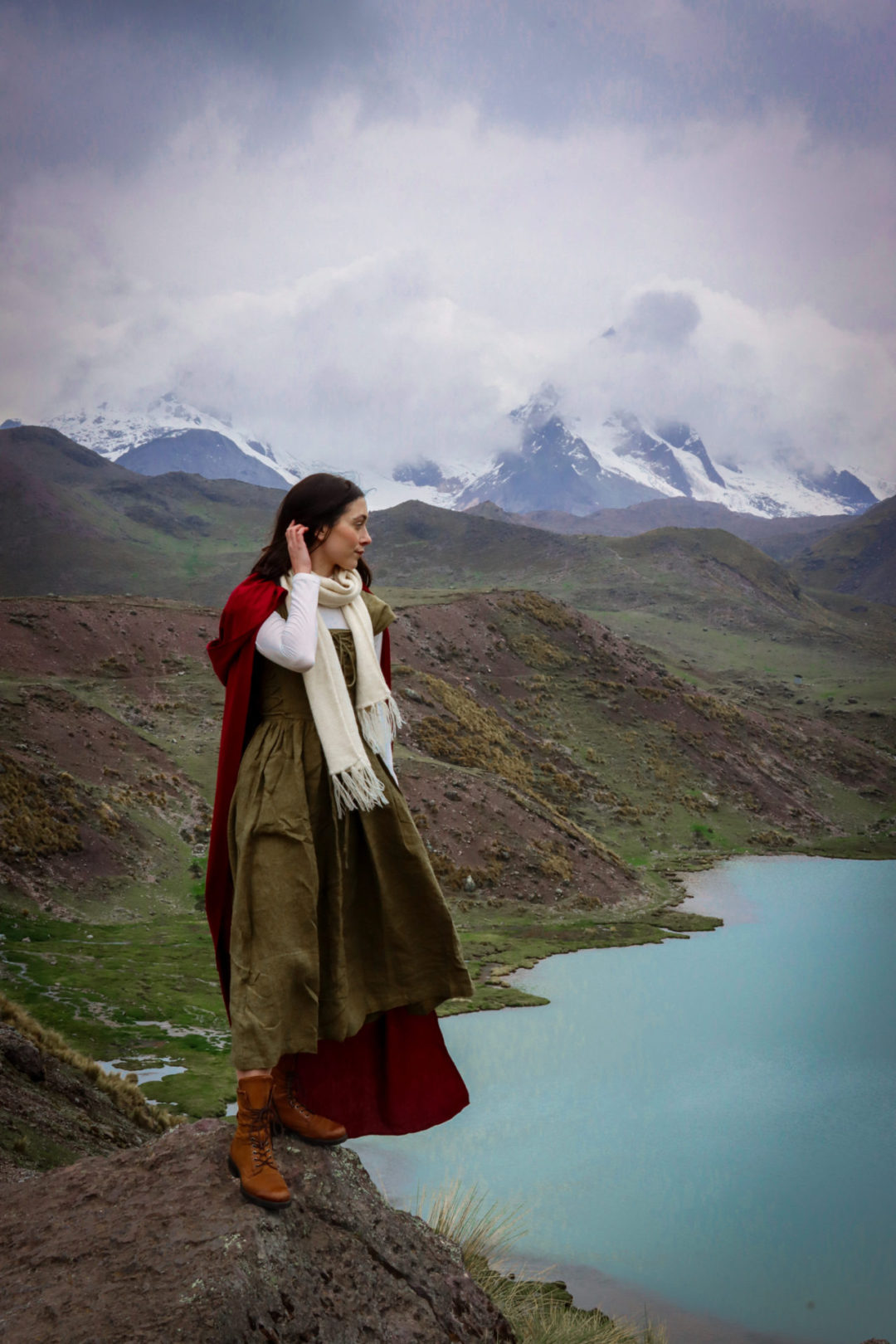 Travel Blogger Jordan Gassner standing on a boulder overlooking  a glacial lake while wearing a dark green fantasy inspired maxi dress, white long sleeve shirt, cream alpaca wool scarf, red cape and brown hiking boots on the Ausangate 7 Lakes Trek near Cusco, Peru