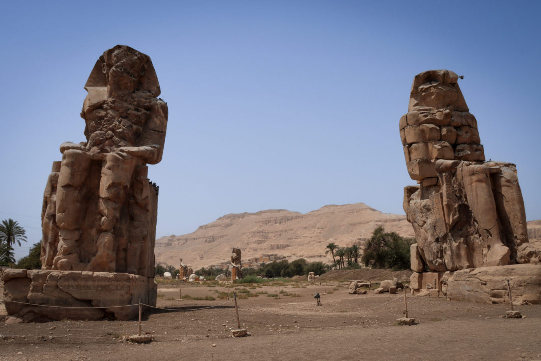 The massive stone statue ruins of Pharaoh Amenhotep III, otherwise known as the Colossi of Memnon, in front of the former grounds of his Mortuary Temple in modern-day Luxor, Egypt