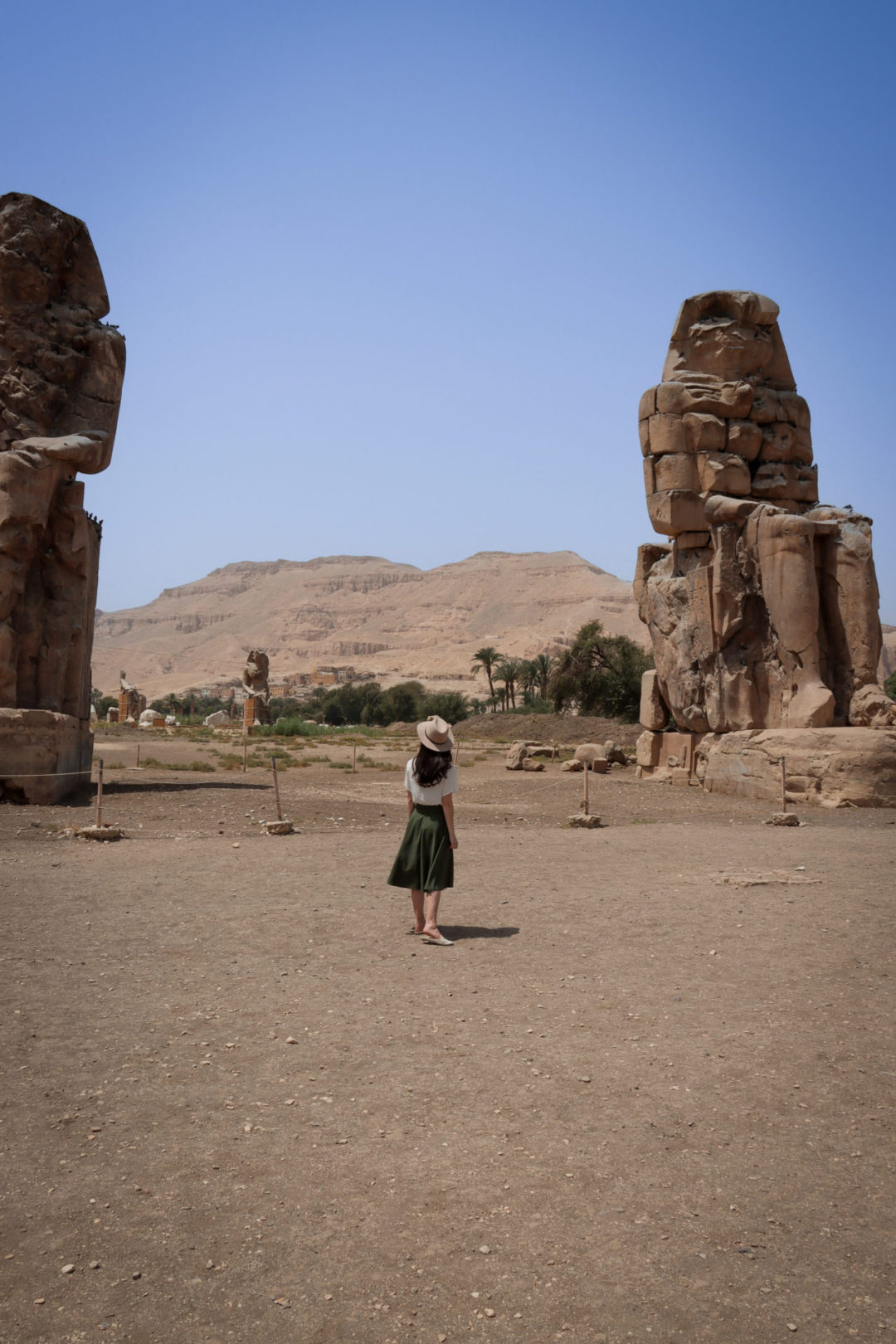 Travel Blogger Jordan Gassner wearing a short sleeve white shirt, army green midi skirt and brown explorer hat standing alone in front of the Colossi of Memnon in Luxor, Egypt