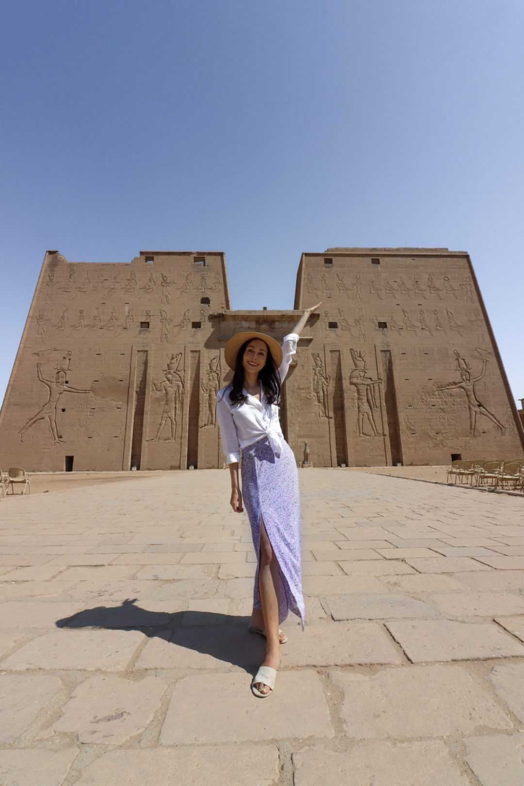 Travel Blogger Jordan Gassner throwing her hand up in the air and smiling while wearing a 3/4-length sleeve white shirt, purple skirt and straw sun hat in front of the entrance to Edfu Temple in Egypt