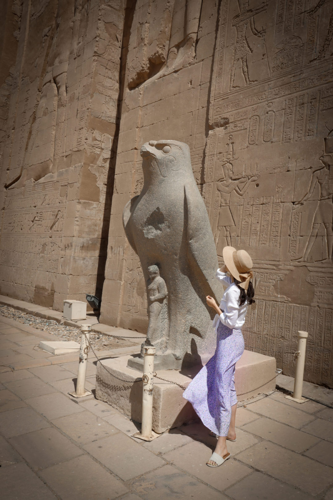 Travel Blogger Jordan Gassner wearing a 3/4-length sleeve white shirt, purple skirt and straw sun hat while looking at a statue of the ancient Egyptian god Horus in front of Edfu Temple in Egypt