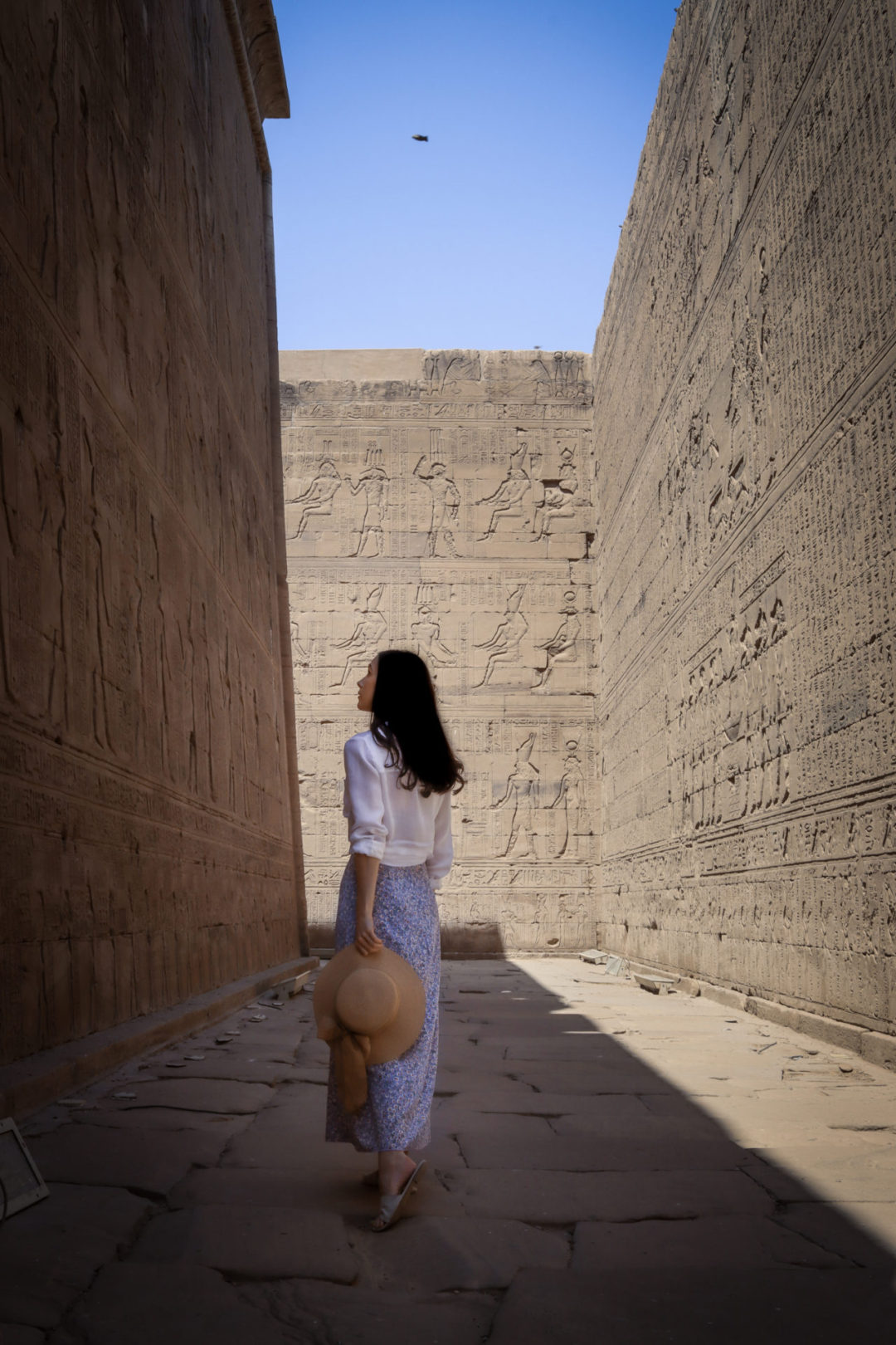 Travel Blogger Jordan Gassner wearing a 3/4-length sleeve white shirt, purple skirt and holding a straw sun hat while walking and looking up at the hieroglyphs dotting the high walls of the Inner Passageway of Edfu Temple in Egypt.