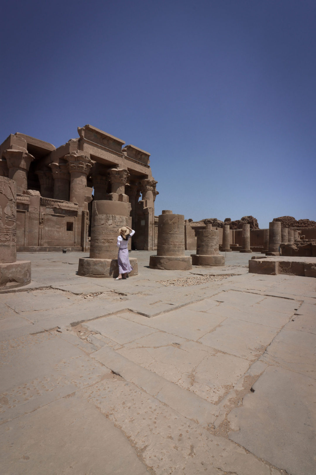 A female traveler holding a straw sunhat to her head and wearing a white shirt and purple skirt reads the heiroglyphs on the remains of a column at the Ancient Egyptian Temple of Kom Ombo