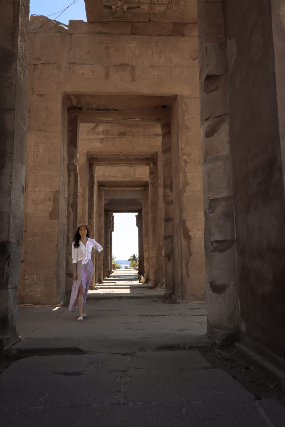 A female traveler wearing a straw, white shirt and purple skirt stands smiling with her left arm on her hip underneath an avenue of arches that leads out toward the Nile River at the Ancient Egyptian Temple of Kom Ombo