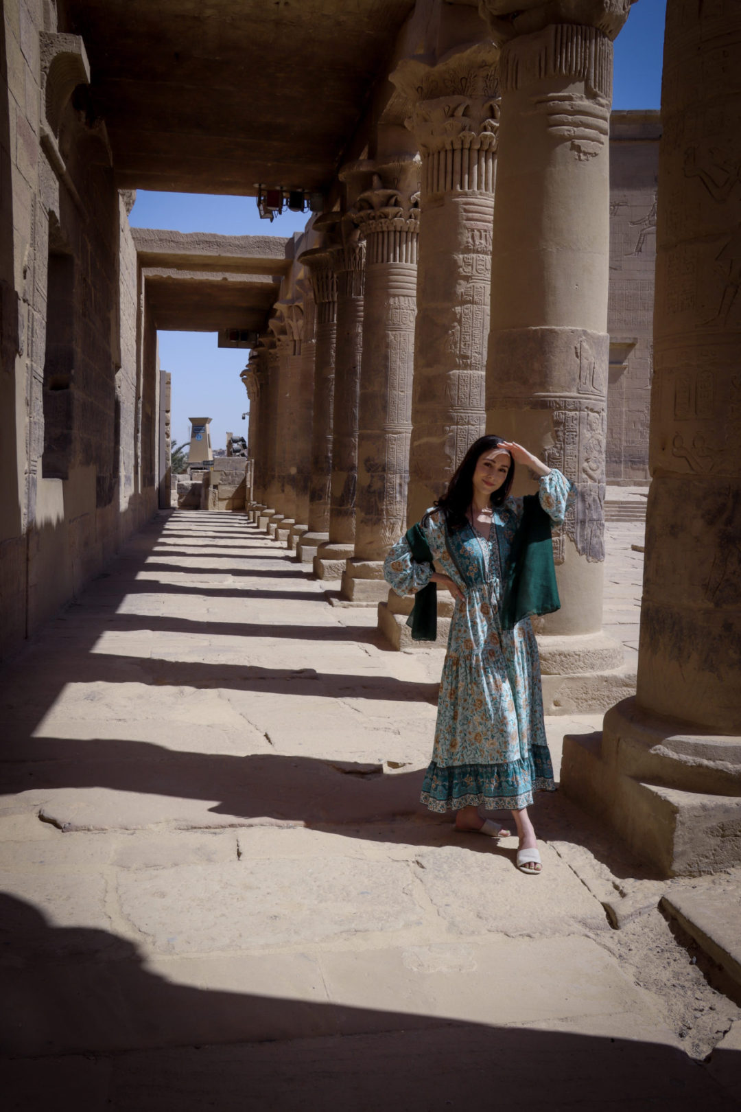 Travel Blogger Jordan Gassner wearing a long sleeve blue patterned maxi dress and a green scarf, shielding her eyes from the sun under a row of columns outside of the Philae Temple ruins on Philae Island in Aswan, Egypt