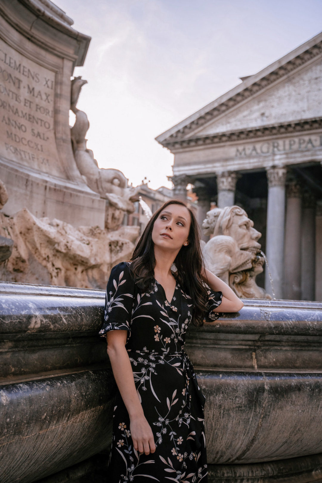 Travel Blogger Jordan Gassner wearing a black floral short sleeve dress while leaning up against an old fountain and touching her hair, peering of into the distance, in front of the Pantheon in Rome, Italy