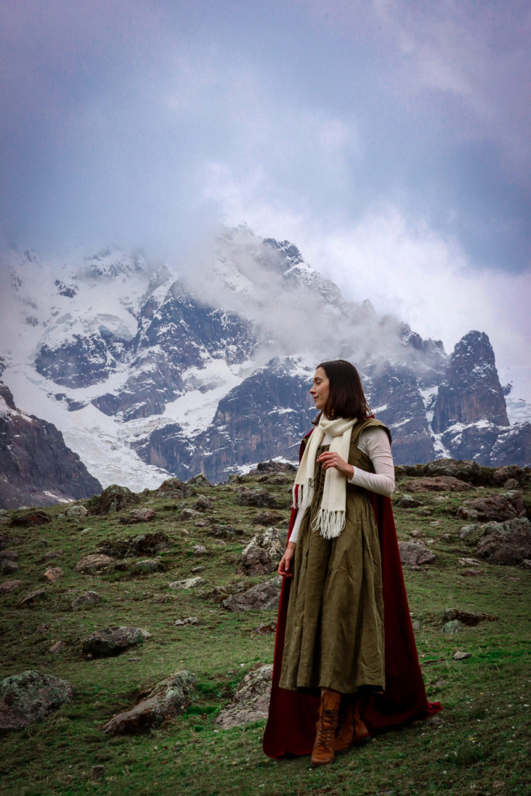 Travel Blogger Jordan Gassner looking off to the side while standing on a hill and wearing a dark green fantasy inspired maxi dress, white long sleeve shirt, cream alpaca wool scarf, red cape and brown hiking boots on the Ausangate 7 Lakes Trek near Cusco, Peru